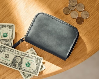 May include: A dark gray leather wallet with a zipper closure, a small wrist strap, and several US dollar bills and coins on a wooden surface. The wallet is rectangular and appears to be new.