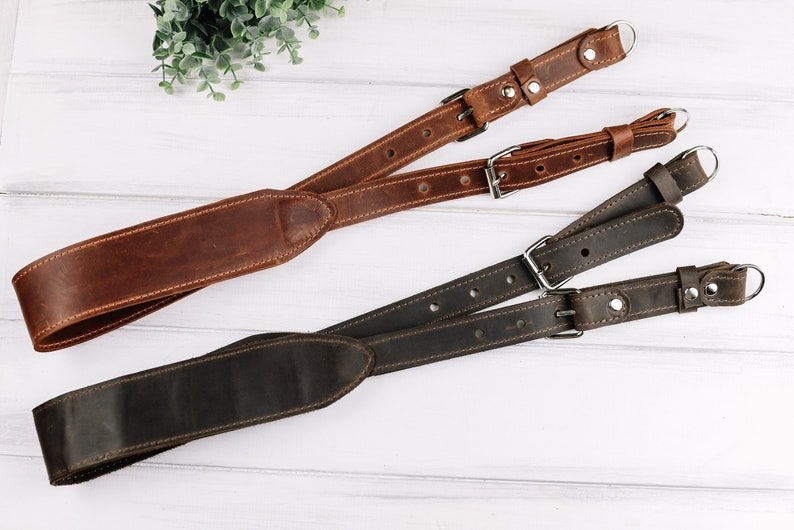 Peut inclure: Deux sangles de cam&eacute;ra en cuir, l'une marron et l'autre noire, avec des boucles et des anneaux m&eacute;talliques. Les sangles sont crois&eacute;es et dispos&eacute;es sur une surface en bois blanc.