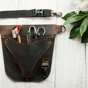 May include: A brown leather tool belt with a black strap and multiple pockets. The belt is filled with gardening tools, including shears, a trowel, and a pair of pruning shears.