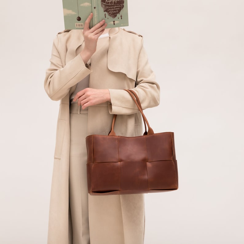 May include: A brown leather tote bag with a woven design. The bag has two handles and is being held by a person wearing a beige coat.