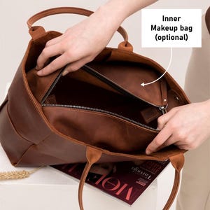 May include: A brown leather tote bag with two top handles. The bag is open and the inside is lined with a brown fabric. There is a zippered pocket inside the bag.