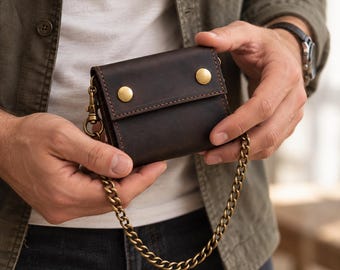 May include: A dark brown leather wallet featuring a fold-over flap secured by two brass-colored metal snap buttons. A matching brass-colored metal chain is attached to the side of the wallet with a swivel lobster clasp hook.