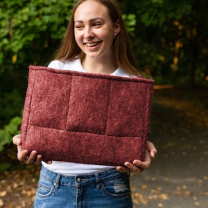 May include: A woman is holding a burgundy felt laptop sleeve with a quilted design. The sleeve is rectangular and made from a soft, textured material.