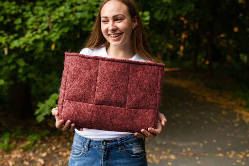May include: A woman is holding a burgundy felt laptop sleeve with a quilted design. The sleeve is rectangular and made from a soft, textured material.