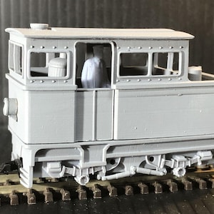 Puede incluir: Una locomotora de tren modelo gris con una figura de una persona en la cabina. La locomotora tiene un diseño detallado con ventanas y un techo. La locomotora está en una vía.