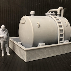 May include: A grey model of an industrial tank with a ladder, pipes, and a small figure of a person standing beside it. The tank is set on a rectangular base with a brick pattern.