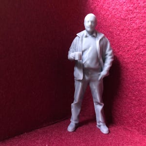 Op de afbeelding: Een grijze miniatuurfiguur van een persoon in vrijetijdskleding, die een mok vasthoudt. De figuur draagt een jas, een overhemd en een broek. De miniatuur staat voor een rode achtergrond.