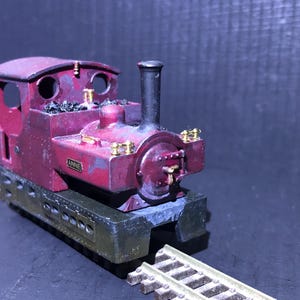 May include: A miniature maroon steam locomotive model with gold accents and the name "ANNIE" on the side. The model sits on a section of model train track. The locomotive has a black smokestack and a dark green base.