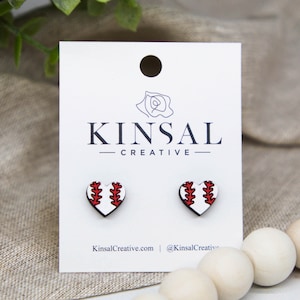 May include: A pair of white heart-shaped earrings with a red baseball design. The earrings are displayed on a white card with the text "Kinsal Creative".