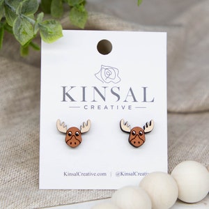 May include: A pair of wooden moose earrings. The moose are brown with white antlers and black eyes. The earrings are on a white card with the text "Kinsal Creative" printed on it.