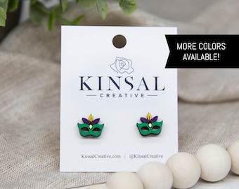 Puede incluir: Un par de pendientes de madera verdes, morados y amarillos con forma de máscaras de carnaval. Los pendientes se muestran en una tarjeta blanca con el texto "Kinsal Creative" y "KinsalCreative.com | @KinsalCreative". El texto "MÁS COLORES DISPONIBLES!" está en la esquina superior derecha de la tarjeta.