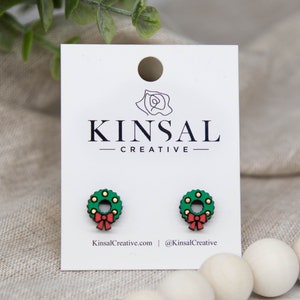 May include: A pair of green and red wooden earrings shaped like a Christmas wreath with a red bow. The earrings are on a white card with the text "Kinsal Creative" printed on it.
