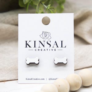 May include: White bone-shaped stud earrings with a dark brown border, displayed on a white card with the text "KINSAL CREATIVE". The card is set against a neutral background with wooden beads.