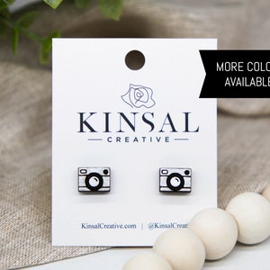 Puede incluir: Un par de pendientes de madera con forma de cámara con un diseño blanco y negro. Los pendientes están sobre una tarjeta blanca con el texto "Kinsal Creative" y "KinsalCreative.com @KinsalCreat". El texto "MÁS COLORES DISPONIBLES!" está en un rectángulo blanco.
