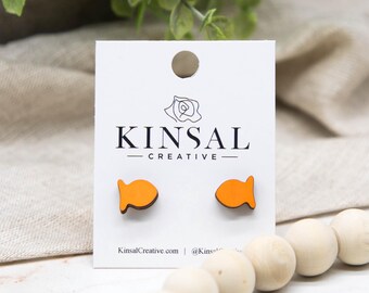 Può includere: Orecchini a forma di pesce arancione su una scheda bianca con la scritta "KINSAL CREATIVE". Gli orecchini sono in legno e hanno un contorno marrone scuro. La scheda include anche il sito web KinsalCreative.com.