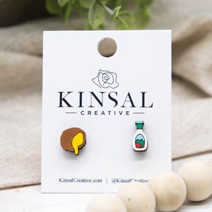 May include: Wooden stud earrings on a white card. One earring is a brown circle with yellow dripping from it, the other is a small bottle with a green top and a red and blue design. The card reads "KINSAL CREATIVE".