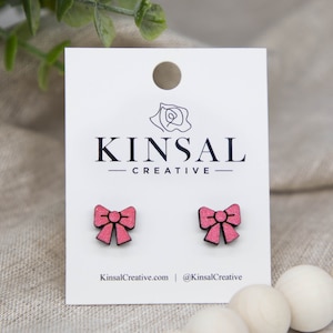 Pode incluir: Brincos de laço rosa em cartão branco com o texto "KINSAL CREATIVE" e endereço do site. Os brincos são de madeira e têm um aspeto texturizado. O fundo é um tecido de linho neutro.