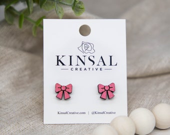 May include: Pink bow-shaped stud earrings on a white card with the text "KINSAL CREATIVE" and the website address. The earrings are made of wood and have a textured appearance. The background is a neutral linen fabric.