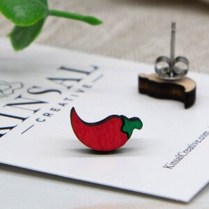 May include: A pair of red chili pepper earrings made of wood. The earrings are on a white card with the text "Kinsal Creative" printed on it.