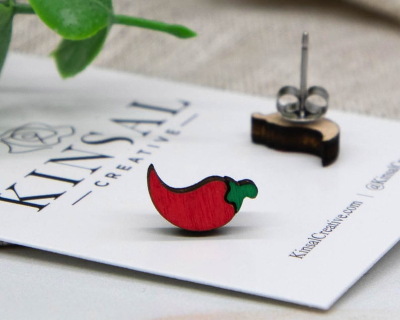 May include: A pair of red chili pepper earrings made of wood. The earrings are on a white card with the text "Kinsal Creative" printed on it.