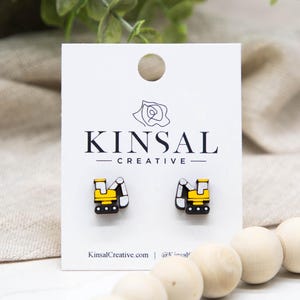 May include: Wooden stud earrings featuring a yellow and black excavator design. The earrings are displayed on a white card with the "KINSAL CREATIVE" logo and website address. The background includes wooden beads and greenery.
