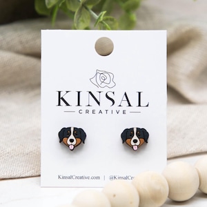 Op de afbeelding: Houten oorstekers met een ontwerp van een Berner Sennenhond. De oorbellen zijn zwart, bruin en wit, met een roze tong. Ze worden gepresenteerd op een witte kaart met de tekst "KINSAL CREATIVE" en de website KinsalCreative.com.