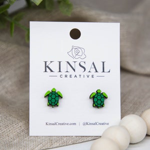 May include: A pair of green and black turtle stud earrings on a white card with the text "Kinsal Creative" printed on it.