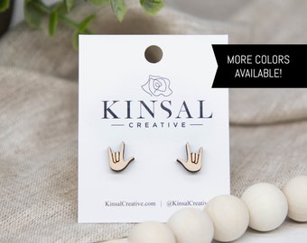 May include: A pair of wooden earrings shaped like hands making the American Sign Language sign for "I love you". The earrings are on a white card with the text "Kinsal Creative" and "KinsalCreative.com | @KinsalCreativ". The text "MORE COLORS AVAILABLE!" is in a black box above the card.