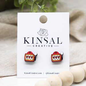 May include: A pair of slot machine earrings with a red, yellow, and black color scheme. The earrings are displayed on a white card with the text "KINSAL CREATIVE" and the website address KinsalCreative.com.