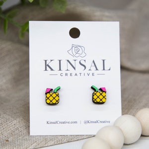 May include: A pair of small, yellow pineapple earrings with green leaves and pink accents. The earrings are on a white card with the text "Kinsal Creative" printed on it.