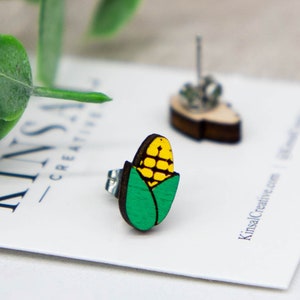 May include: A pair of wooden earrings shaped like corn on the cob. The corn is yellow with green husks. The earrings are on a white card with the text "Kinsal Creative" printed on it.