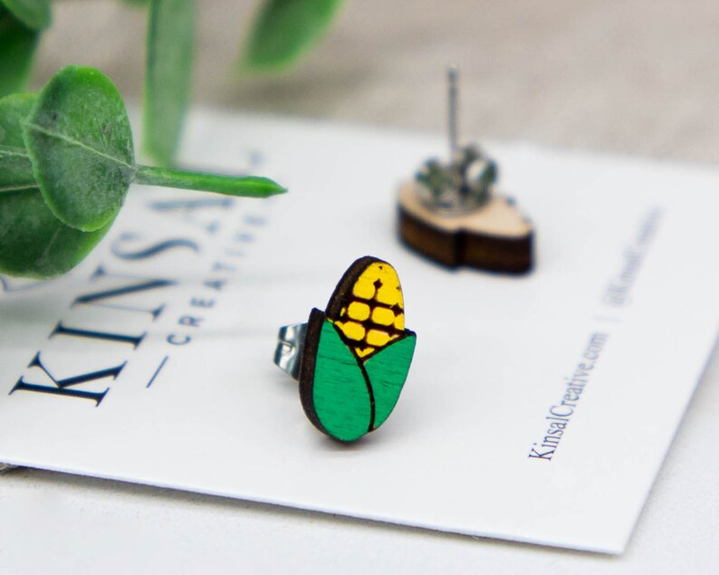May include: A pair of wooden earrings shaped like corn on the cob. The corn is yellow with green husks. The earrings are on a white card with the text "Kinsal Creative" printed on it.