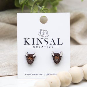 May include: Wooden stud earrings featuring a bison design. The earrings are brown with white horns and a detailed face. The earrings are displayed on a white card with the text "KINSAL CREATIVE" and the website KinsalCreative.com.