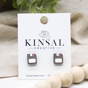 Puede incluir: Pendientes de madera con forma de disquetes en miniatura. Cada pendiente presenta un cuadrado gris, un rectángulo blanco y un acento rojo. Se exhiben en una tarjeta blanca con el texto "KINSAL CREATIVE".