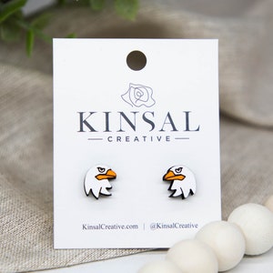 May include: A pair of white wooden earrings shaped like the head of an eagle with orange beaks. The earrings are on a white card with the text "Kinsal Creative" printed on it.