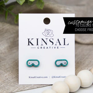 May include: Wooden stud earrings in the shape of goggles, featuring a silver glitter centre and teal edging. The earrings are presented on a white card with the text "customize me 70 COLORS TO CHOOSE FROM!" and the brand name "KINSAL CREATIVE".