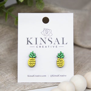 以下が含まれることがあります： 緑の葉と黄色の果実が付いた木製のパイナップルイヤリングのペア。イヤリングは、「Kinsal Creative」というテキストが書かれた白いカードに表示されています。