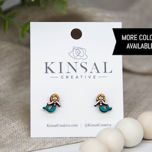 May include: A pair of wooden mermaid stud earrings with a teal tail, pink top, and blonde hair. The earrings are displayed on a white card with the text "Kinsal Creative" and "More Colors Available!"