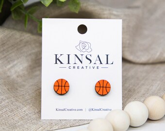 May include: A pair of orange and black basketball stud earrings on a white card with the text "Kinsal Creative" and the website address "KinsalCreative.com" and the social media handle "@KinsalCreative".