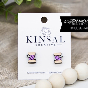 May include: A pair of wooden earrings shaped like spools of thread with a needle sticking out. The thread is pink and the spools are a light brown color. The earrings are on a white card with the text "Kinsal Creative" and "customize me 70 colors to choose from!"