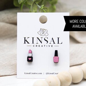 May include: Wooden stud earrings on a white card. One earring is a pink lipstick with a black base and a heart. The other is a pink nail polish bottle with a black cap. The card has the text "KINSAL CREATIVE" and "MORE COLORS AVAILABLE!"