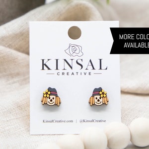 May include: A pair of wooden earrings featuring a scarecrow design. The earrings are painted in shades of brown, yellow, and red. The earrings are displayed on a white card with the text "Kinsal Creative" and "More Colors Available!" printed on it.