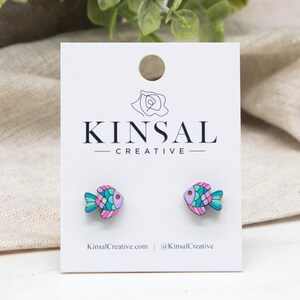 May include: A pair of small stud earrings shaped like fish, featuring scales painted in vibrant shades of pink, purple, and teal. The earrings are mounted on a white display card printed with the text KINSAL CREATIVE and KinsalCreative.com.