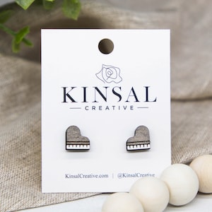 May include: A pair of wooden earrings shaped like a piano. The earrings are gray with white keys. The earrings are on a white card with the text "Kinsal Creative" printed on it.