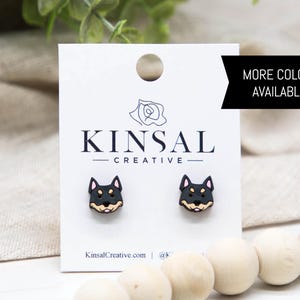 Könnte beinhalten: Ohrstecker mit Shiba Inu-Gesicht in Schwarz und Hellbraun auf einer weißen Karte. Die Karte trägt den Text "KINSAL CREATIVE" und ein schwarzes Banner mit der Aufschrift "MORE COLORS AVAILABLE!". Die Ohrringe werden auf einer beigen Oberfläche mit Holzkugeln präsentiert.