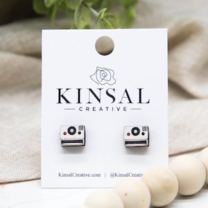 May include: A pair of wooden Polaroid camera stud earrings on a white display card. The earrings are small, square-shaped, and feature a black and white design with a red dot. The card has the text "KINSAL CREATIVE" and a website address.