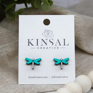 Op de afbeelding: Een paar turquoise blauwe libellen oorbellen op een witte kaart met de tekst "Kinsal Creative" erop gedrukt.