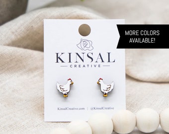 May include: A pair of white chicken stud earrings with red combs and yellow feet. The earrings are on a white card with the text "Kinsal Creative" and "More Colors Available!" printed on it.