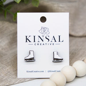 May include: A pair of white wooden ice skate earrings. The earrings are on a white card with the text "Kinsal Creative" printed on it.
