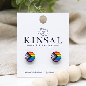 May include: Round wooden stud earrings with the Progress Pride flag design, displayed on a white card. The earrings feature a rainbow colour scheme with black and brown accents. The card has the "Kinsal Creative" logo and website address. The earrings are a small accessory.